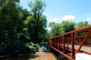 A pedestrian bridge with red metal trusses on both sides and wooden decking on the walkway stretches across a brown creek or river. There are tall trees on the opposite shore.