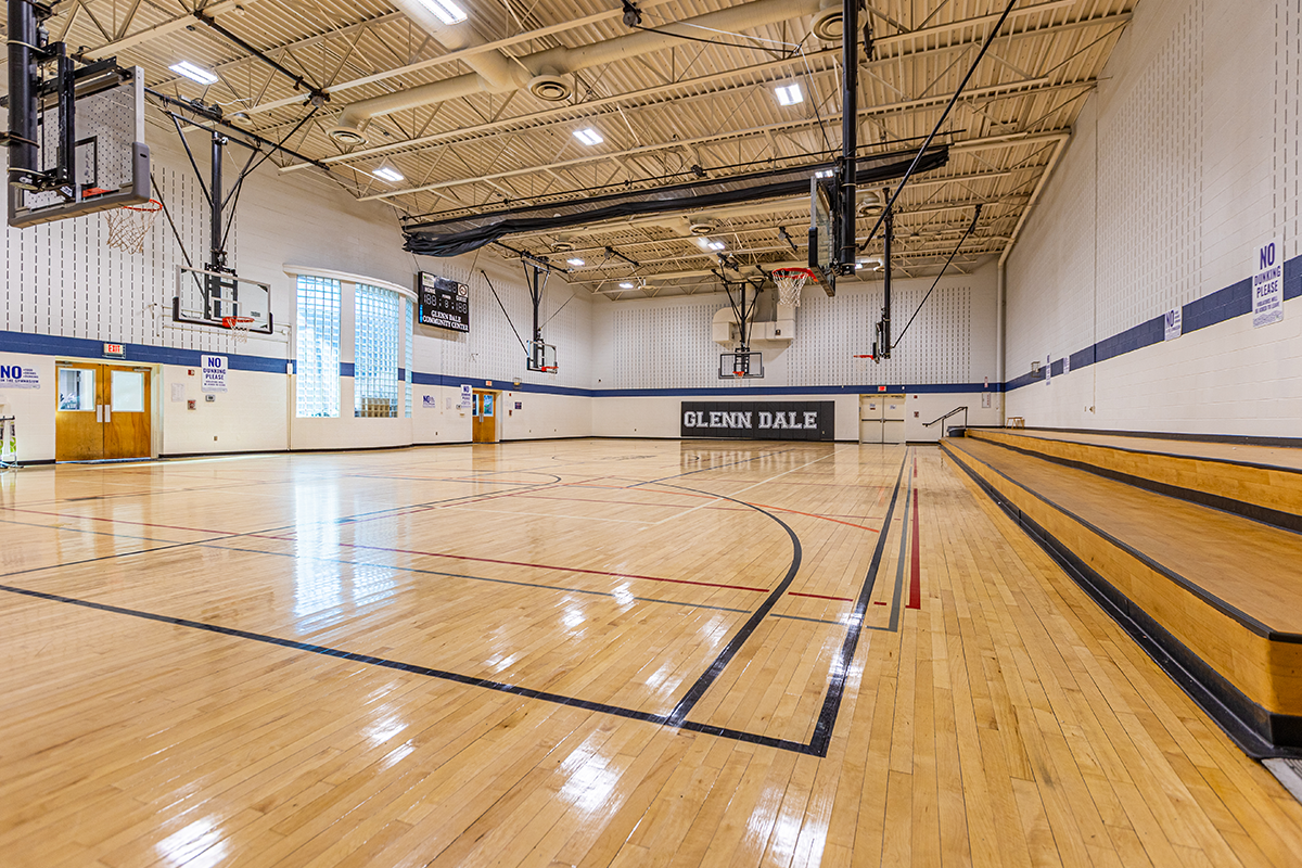 A large gymnasium with multiple basketball goals, wooden bleachers down one wall, and a large, curved, glass-block window in the opposite wall, next to a scoreboard. On the far wall are the words GLENN DALE.