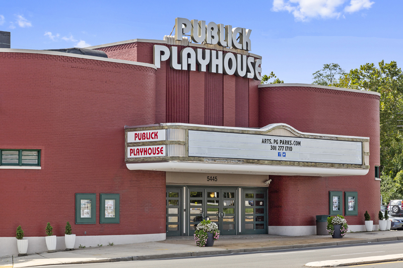 Exterior view of a large, art deco, brick theatre painted red, with a big white marquee sign over the entrance, and the words PUBLICK PLAYHOUSE on the top in white art deco letters. The front of the building has rounded corners, curving to the side walls on either side of the marquee. Several potted plants are on the sidewalk out front.