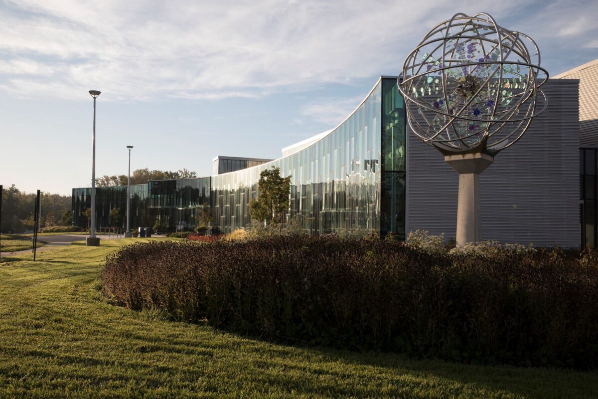 A large, glass and concrete building. Next to the corner of the building is a tall, spherical sculpture on a post.