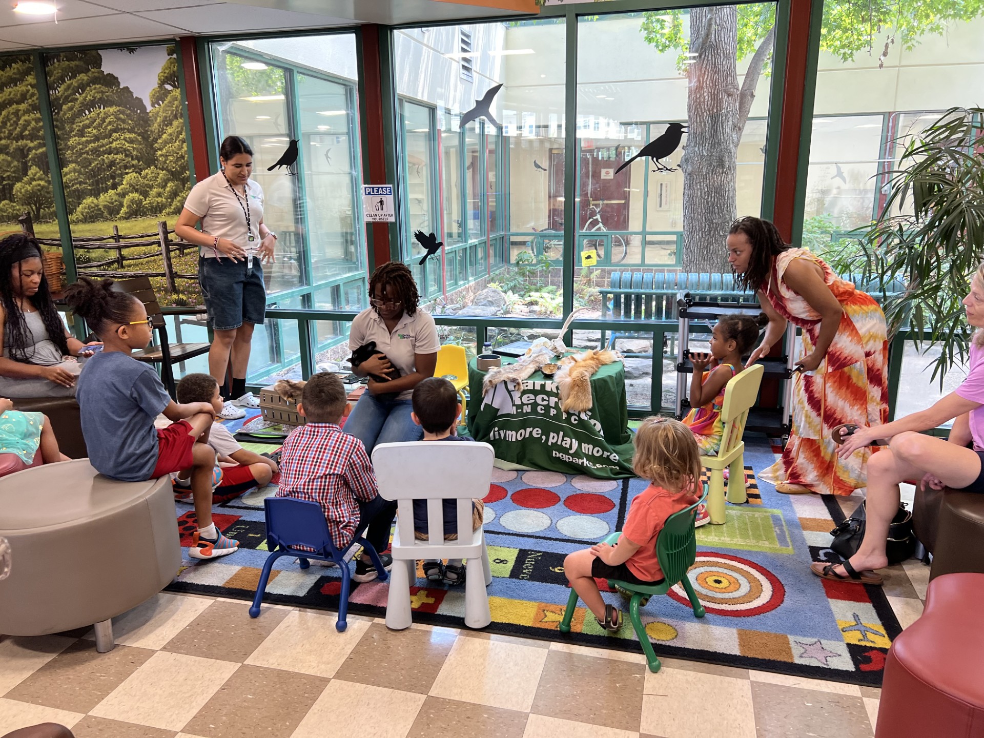 A staff member kneels on a colorful carpet holding a black rabbit, surrounded by children.