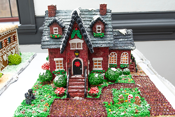 A red brick gingerbread house. It has icing-lined shingles, pretzel window panes, frosting grass, peanut butter cup bushes and a driveway and walkway made out of cobblestone coffee beans