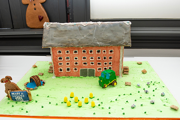 A large gingerbread building with a frosted roof. There are many icing-lined windows facing he front of the property, where there is a tractor, a water feature, and some haybales made of mini wheats.