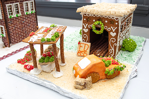 A beautifully decorated gingerbread farmers market building, with frosting and winter decorations lining the sides and roof made of Rice Chex. The pretzel market stall is ready to sell all sorts of goods to gingerbread patrons.