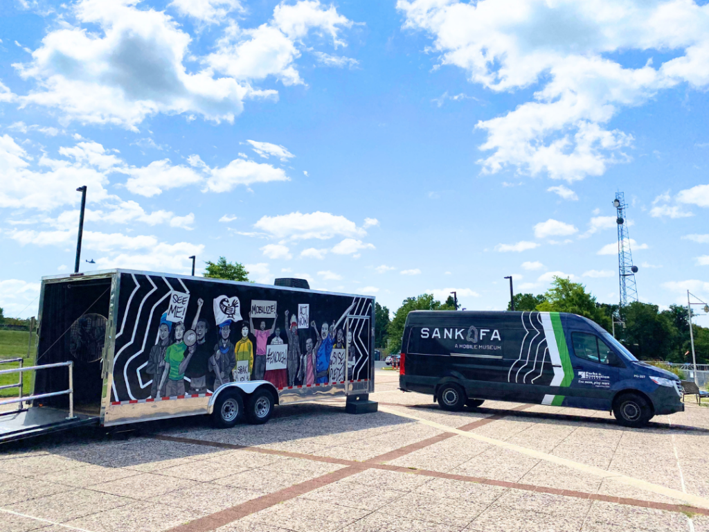 A van with the word SANKOFA on it is parked next to a large trailer decorated in a colorful mural.