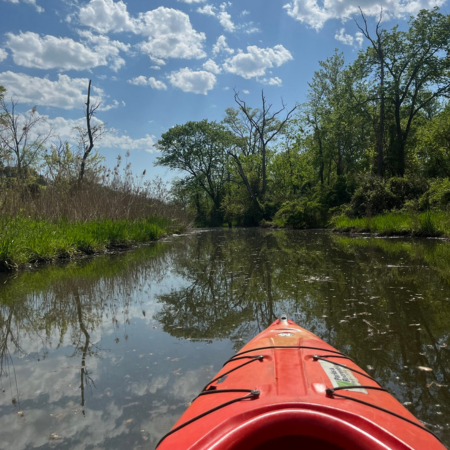 A calm inlet or river seen from the perspective of a person in a red plastic kayak. The water is very still and reflects clouds.