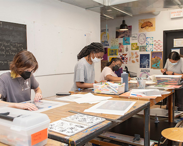 In a classroom, four young women wearing masks work at large wooden tables on art projects. There are several dozen artworks hanging on the far wall.