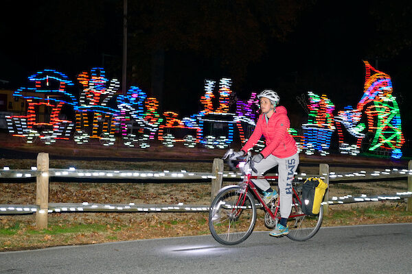 A woman on a bike in a red sweater is riding past a holiday light decoration shaped like a train.