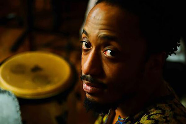A man wearing a colorful, patterned tunic raises his eyebrows. Next to him is a traditional drum.