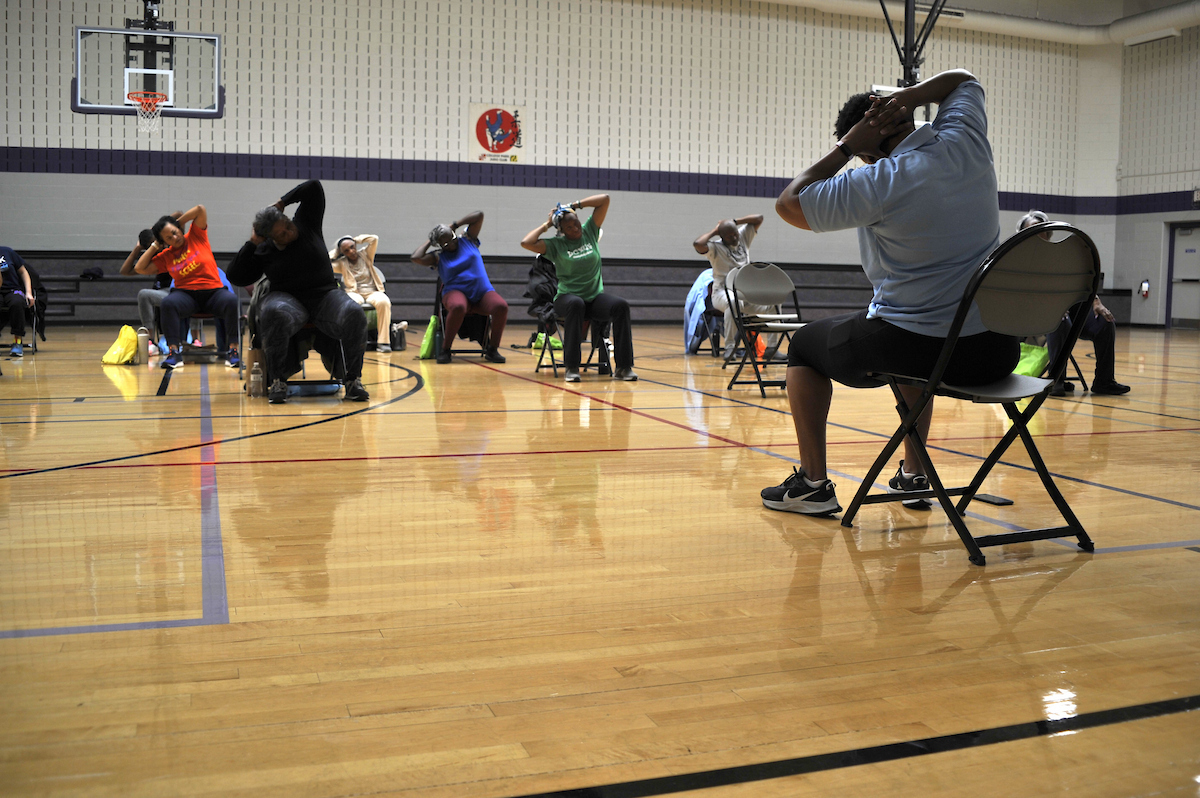 In a gymnasium, a class of adults sitting in folding chairs does a stretch to the side with their arms behind their heads. In front of the class, an instructor in a light blue shirt demonstrates the stretch.