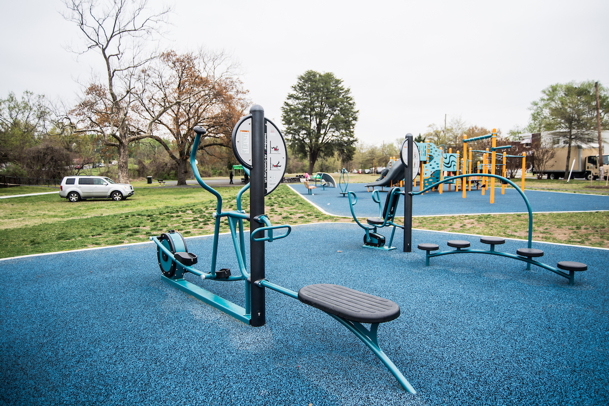 Several pieces of outdoor fitness equipment, including an elliptical trainer, in a park. Behind the fitness area there is a blue and yellow playground.