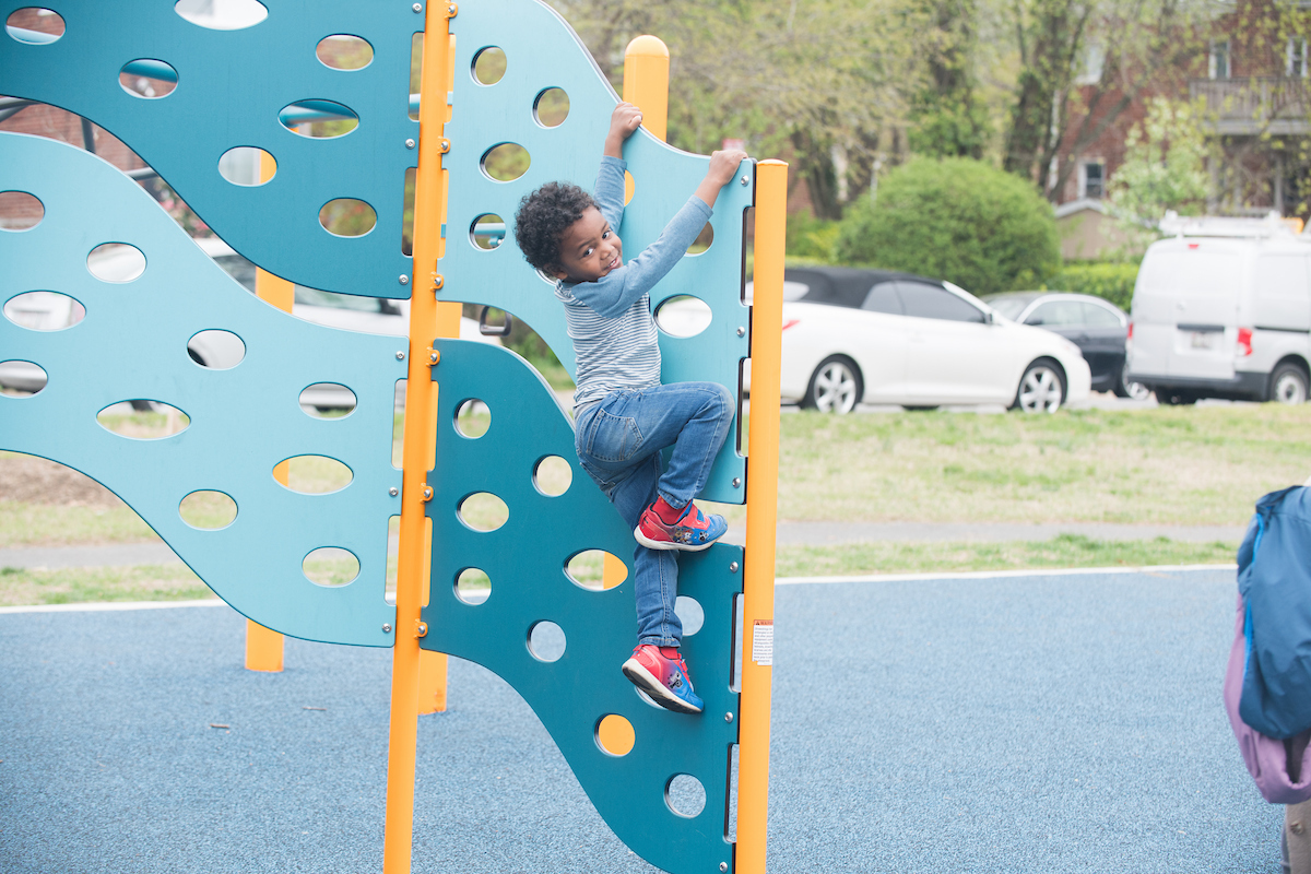 A child climbs on a vertical, blue climbing panel at an outdoor playground. The panel is wavy and filled with holes like Swiss cheese, for hand and foot-holds.