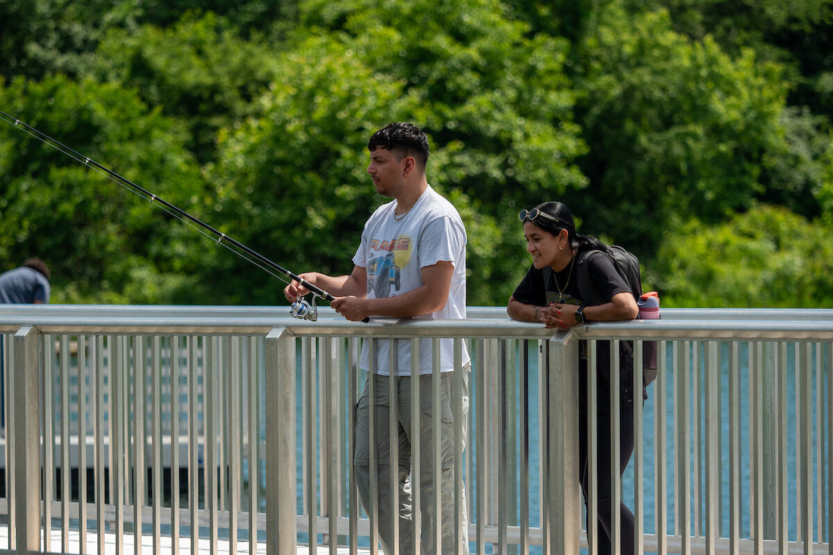 A man fishes off the side of a pier, while a woman at his side watches. It's a sunny day, and the water reflects the blue sky.