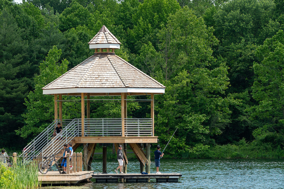 A large, octagonal, wooden gazebo on a low wooden pier that juts out into a lake. The gazebo is elevated off the pier, with a wooden staircase leading up to the gazebo. A boy fishes off the pier. Nearby, another boy stands resting near his bicycle. Across the water are green trees.