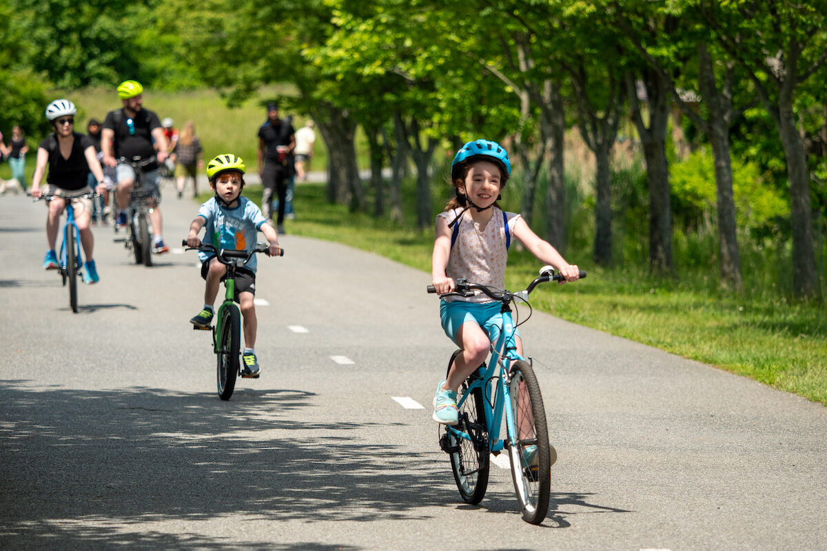 Two small children and their parents ride bikes on an asphalt trail with a dotted white line down the middle. There are people walking and jogging on the trail in the background, and someone walking a dog. The cyclist family all wear helmets that match the color of their bicycles.