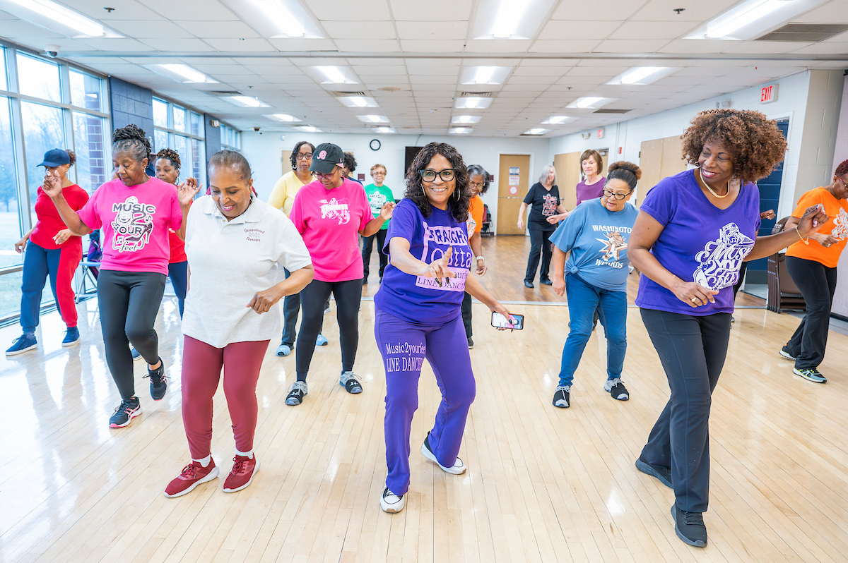 In a dance studio with one mirrored wall and bank of floor-to-ceiling windows, about a dozen smiling women perform a line dance in formation. Many are wearing colorful t-shirts with the names of line dancing groups on them, such as "Music 2 Your Feet Line Dancing."