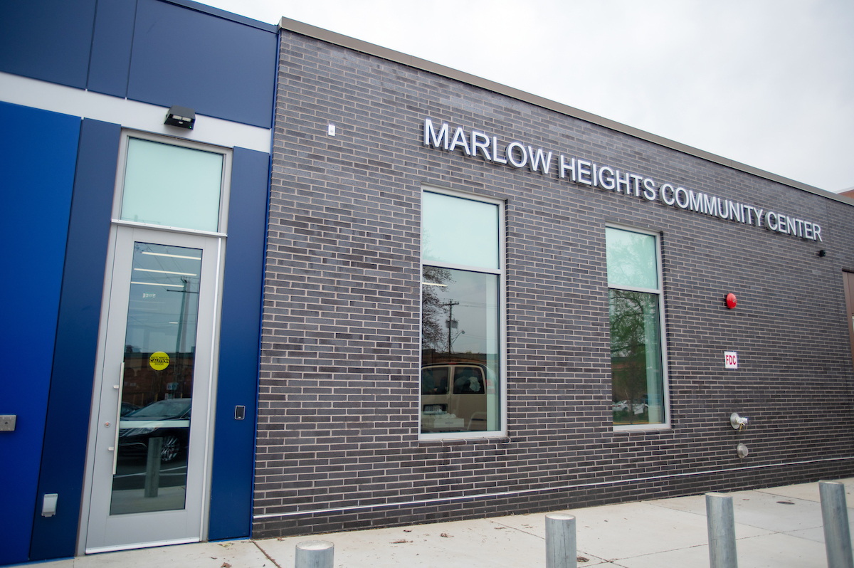 Close-up view of the front facade of a large building that is partially made of gray brick and partially clad in blue metal panels, with glass windows and a door. Letters on the building spell MARLOW HEIGHTS COMMUNITY CENTER.