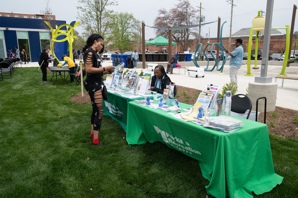 In the grass in front of a large, modern building, two long information tables are set up and piled with literature and flyers. To the right rear is a large, concrete splash pad with various colorful pieces of equipment and play areas, and a shade sail. A woman wearing black stands at one table reading a flyer.