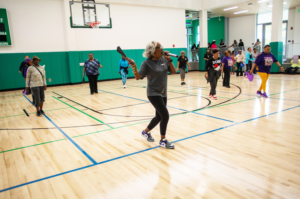 Inside a gymnasium with a wooden floor and green padded walls, a class of adults stands in formation doing aerobic moves. The instructor is positioned in the front of a class, holding a microphone.