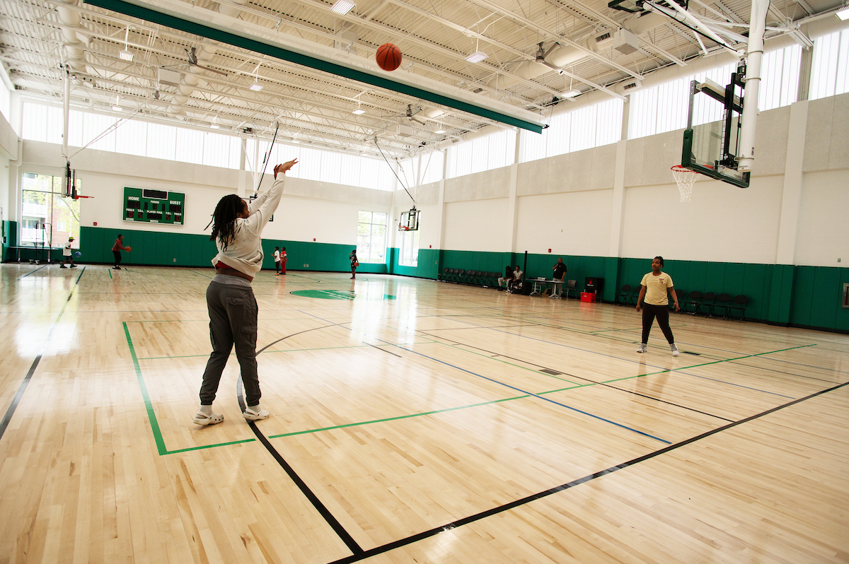 Inside a large gymnasium with wooden floors and green padded walls, two women play basketball. The entire top section of the gymnasium's walls is made of windows. A green scoreboard hangs on the far wall.