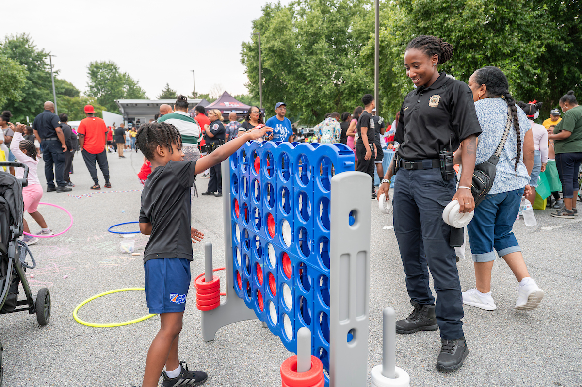 In a parking lot, a boy plays oversized Connect Four against a woman wearing a park police uniform and an amused expression. Behind them, kids play with hula hoops and jump ropes. There are adults crowded along the edge of the parking lot, where long information tables and vendors are set up. At the far end of the parking lot is a temporary bandshell.