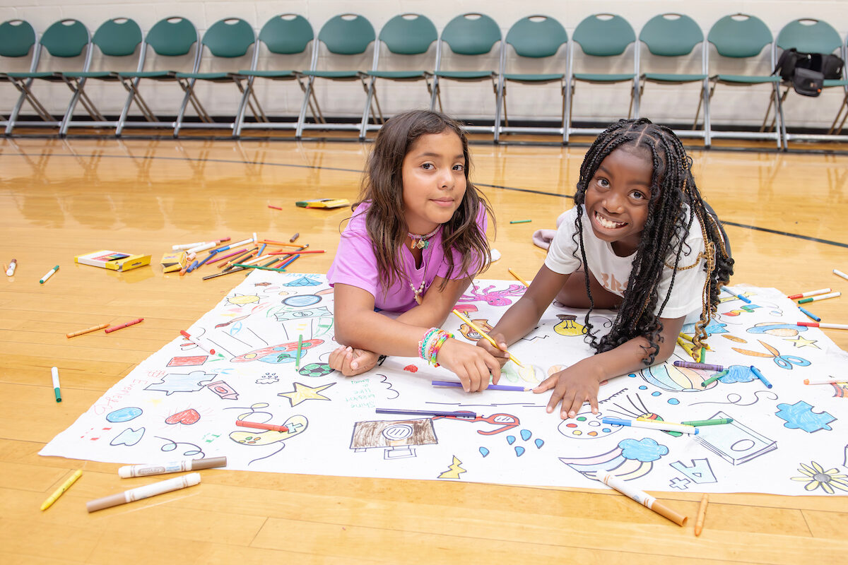 Two smiling girls are sprawled out on the floor of a gymnasium working on a large paper banner that they are coloring with markers. On the floor all around them are scattered dozens of colorful markers and colored pencils.