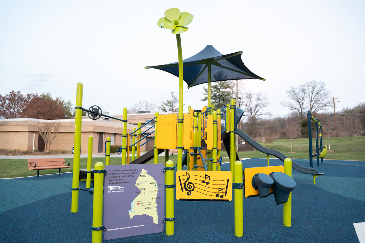 A green and blue outdoor playground with multiple slides, platforms, and climbing features, in the grass outside a beige, brick building. The playground features musical instrument elements as well as a panel containing a map of Prince George's County with several other notable playgrounds marked on the map.