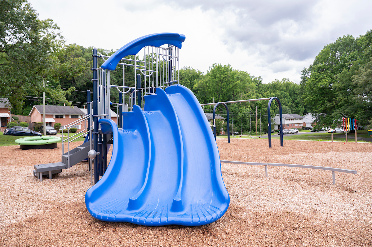 A blue play structure at an outdoor playground featuring three blue plastic slides of increasing heights, along with stairs, platforms, and monkey bars. Behind this structure are swings, xylophones, a balance beam, and a spinning ring feature.