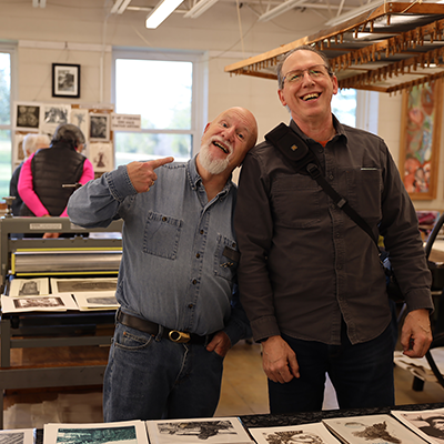 Two smiling men pose in an art studio, with one leaning his head on the other's shoulder and pointing up at him. In front of them on a low table are half a dozen small prints in black and blue ink. Behind them is a printmaking press. To the right, a rack hangs from the ceiling with dozens of clothespins attached to it.