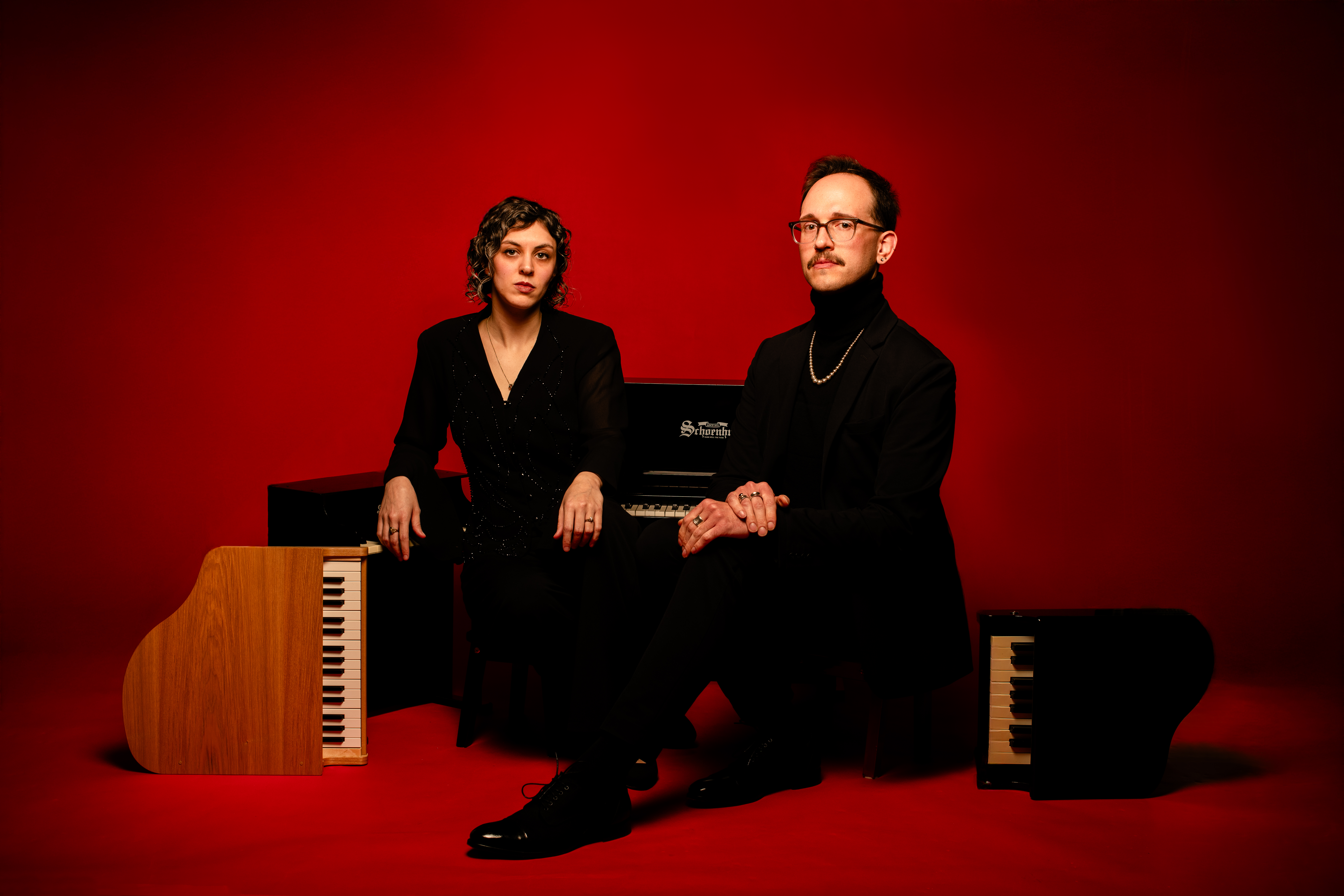 Studio portrait of two people in black attire seated at keyboard instruments against a solid red background.