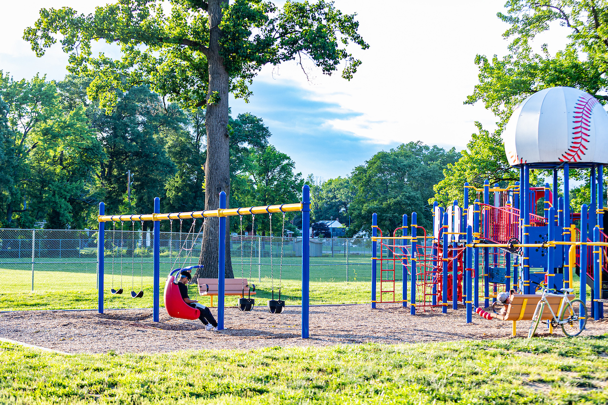 Someone sits in the accessible swing on a blue swingset at an outdoor playground on a sunny day. Nearby is a large play structure featuring rope webs, platforms, bridges, slides, climbing elements, and more. The central tower is topped with a roof that looks like a giant baseball.