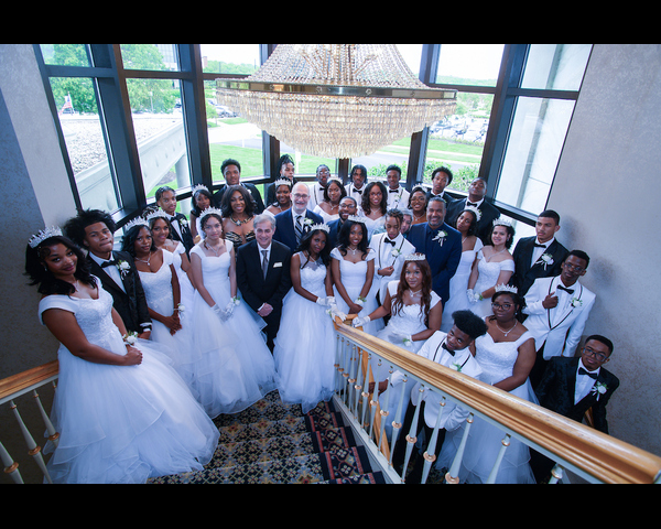 Several dozen people in formalwear pose on the landing of a large, carpeted stairwell with an ornate crystal chandelier above them, and a massive window behind them. Young women are all wearing white gowns and tiaras; young men are all wearing tuxedos. They all have corsages and boutonnieres with white flowers.