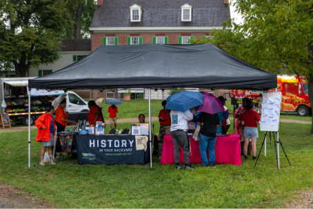 Visitors gather beneath a black canopy tent featuring a local history display during an outdoor community event.