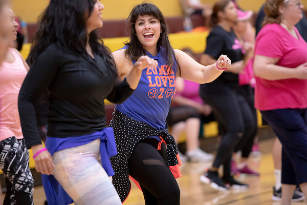A class of women wearing workout clothes exercises in a gymnasium. The woman in the middle of the photo is smiling and wearing a blue shirt with the words ZUMBA LOVE printed on it.