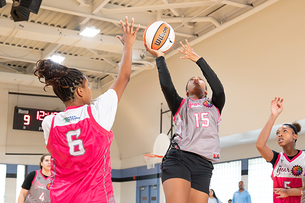 Girls playing basketball. A girl in a red jersey with braids defends her hoop against a girl in a grey jersey as she attempts a layup.