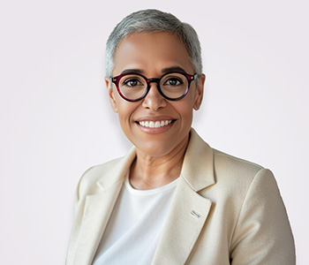 A smiling woman with short hair wearing a beige suit jacket and red glasses.