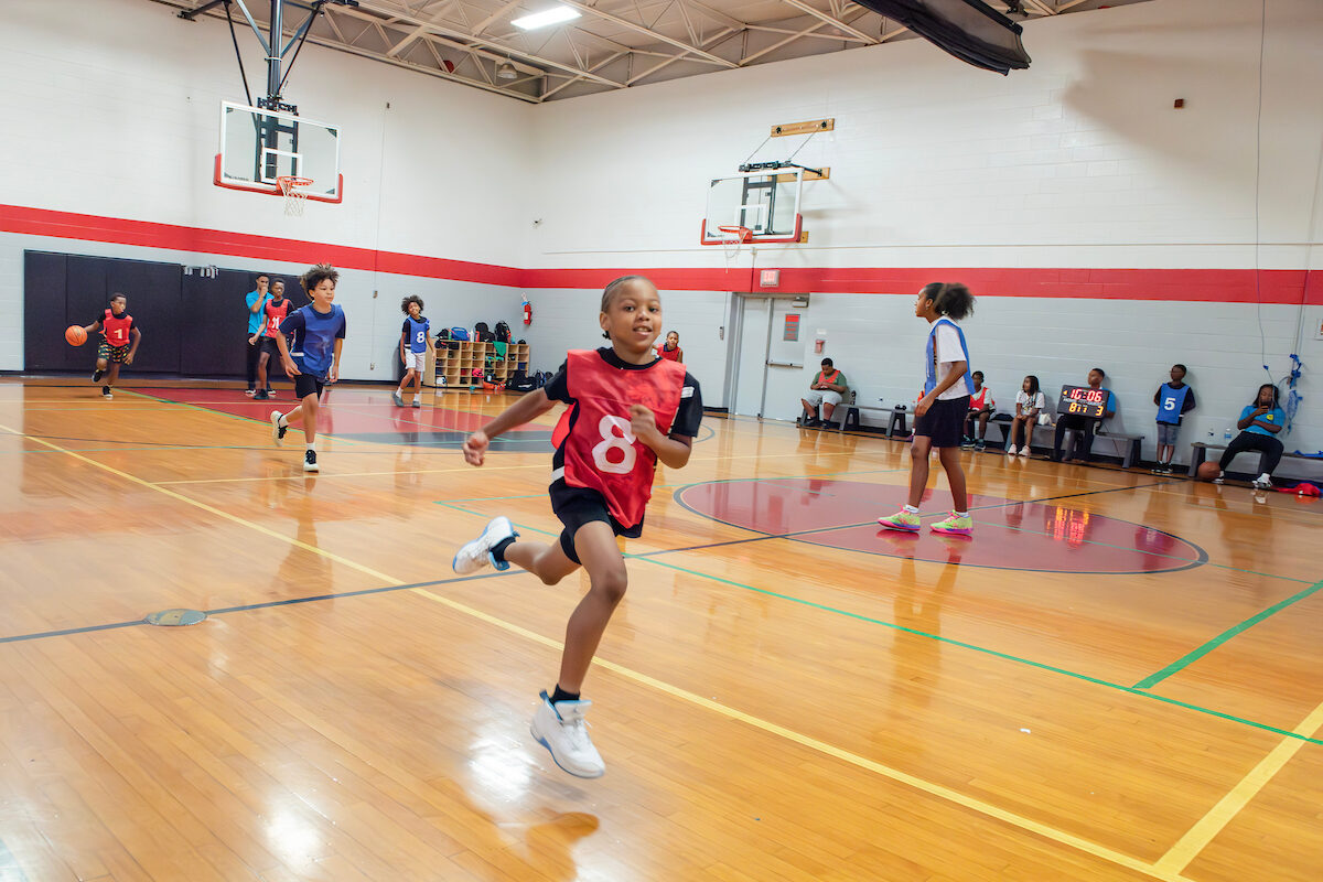 Boys in red and blue numbered smocks play basketball in a gymnasium.