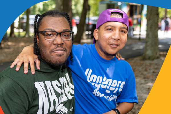 Two young men pose in a park with their arms around one another's shoulders.