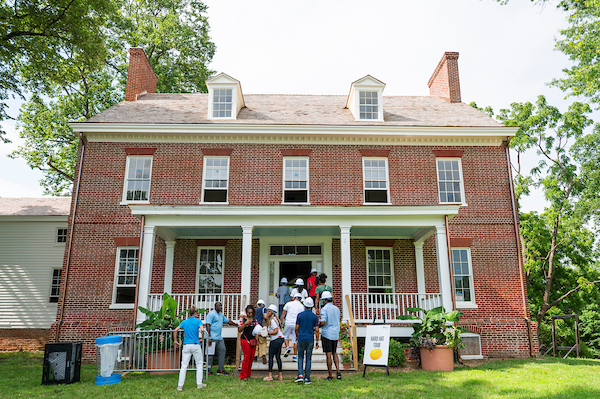 About 20 people in hard hats climb the porch steps to enter a two-story, Colonial brick building. A sign in the grass reads HARD HAT TOUR.