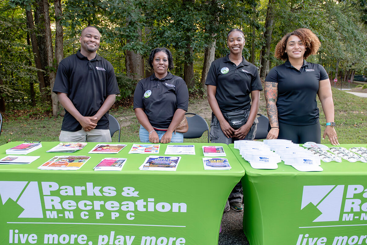 Four smiling staff members stand behind two information tables set up in the grass in a park. The tables contain flyers and giveaway items.