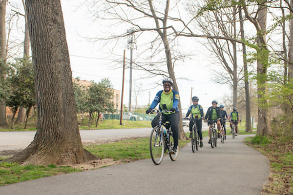 Five people in matching green and blue cycling jerseys ride bikes in a single file line on a flat, paved trail.