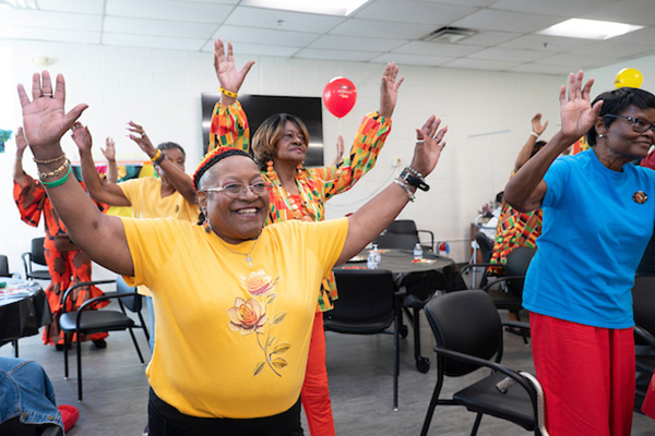 Half a dozen smiling seniors dance with their arms raised in a multipurpose room decorated with balloons.