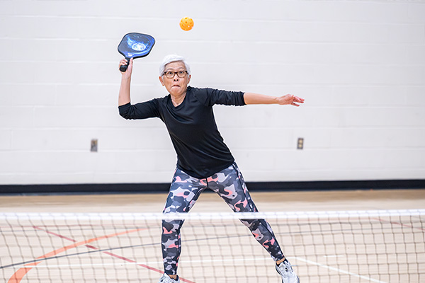 A senior woman prepares to his a pickleball with a blue paddle on an indoor pickleball court.