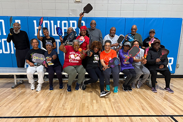 About 16 smiling adults pose holding pickleball paddles in a gym. Blue mats on the wall behind them have white lettering that reads John E. Howard Community Center.