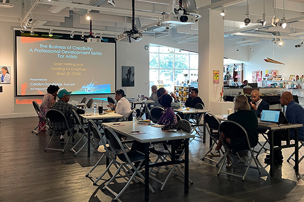 People sit at tables set up in an art gallery with a slide presentation on the far wall titled, "The Business of Creativity."