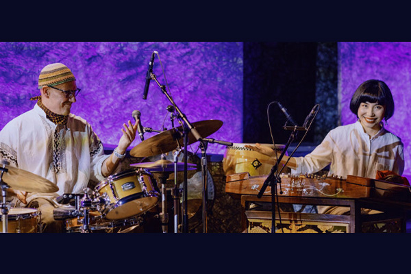 A musical performance by a man sitting behind a drum kit and a woman sitting behind a large Chinese dulcimer.