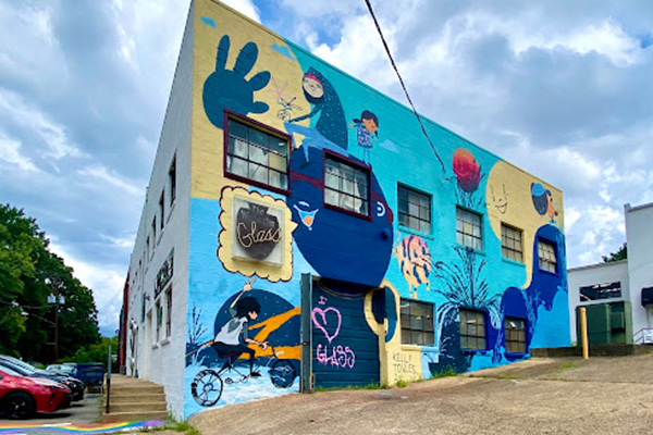 A two-story cinderblock building with a colorful mural covering one whole side. A black garage door is spraypainted with "I heart Glass."