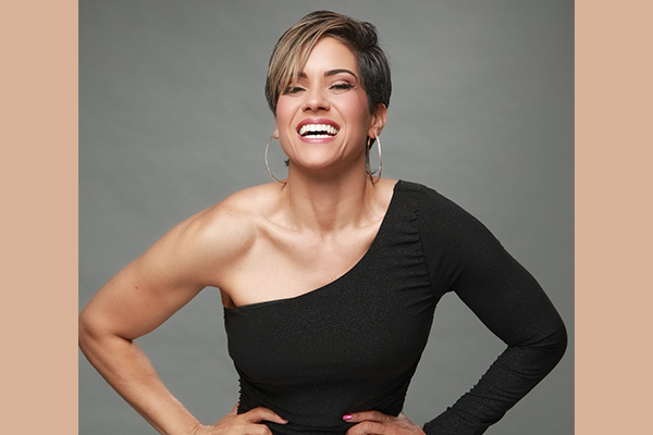 Headshot of a smiling woman with her hands on her hips, wearing a one-sleeved black top.