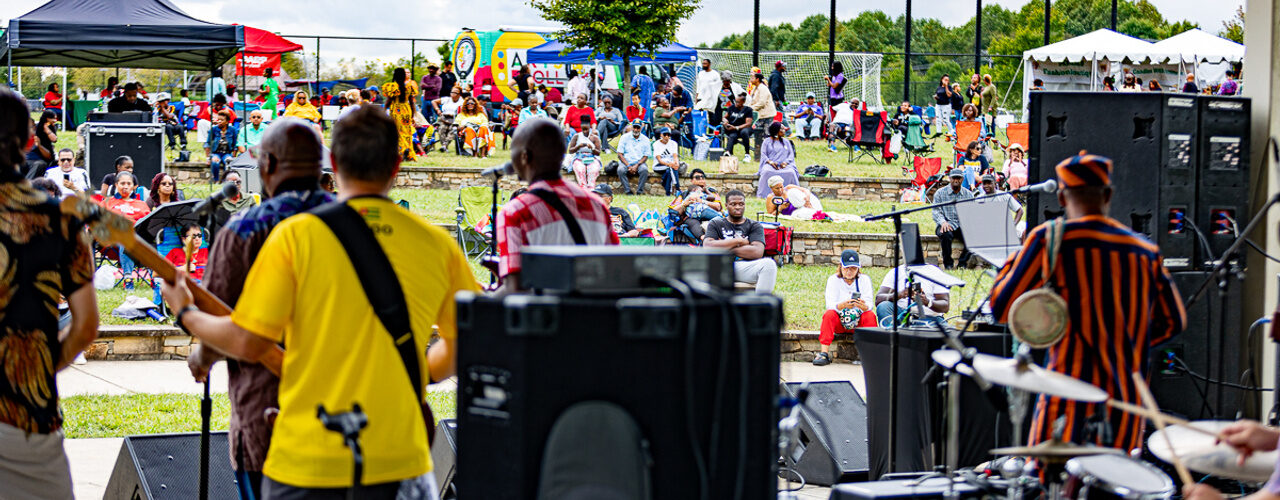 View from the stage of musicians performing for a large outdoor festival audience seated and standing on a grassy hillside.