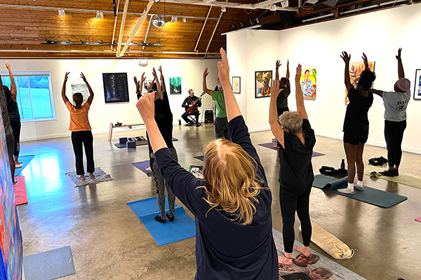 Inside an art gallery, adults stand on yoga mats being led in a stretch by an instructor. Next to the instructor is a seated man playing the guitar.